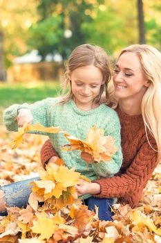 Happy young caucasian mother and little daughter holding autumn yellow leaves sitting at the park