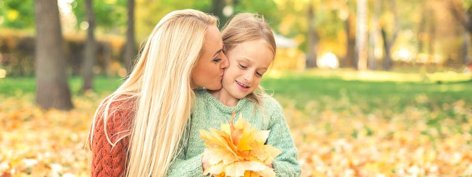 Happy young caucasian mother and little daughter holding autumn yellow leaves sitting and kissing at the park