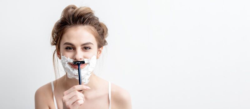 Beautiful young caucasian woman shaving her face by razor on white background. Pretty woman with shaving foam on her face