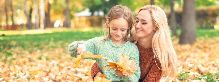 Happy young caucasian mother and little daughter holding autumn yellow leaves sitting at the park