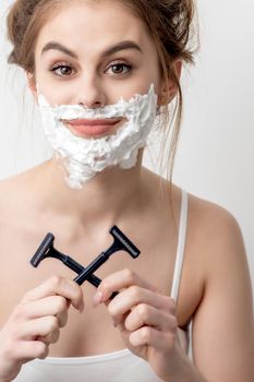 Beautiful young caucasian woman with shaving foam on her face and two razors in her hands on white background