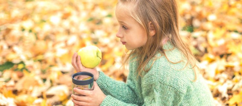 Beautiful little caucasian girl with apple and drink sitting in autumn park