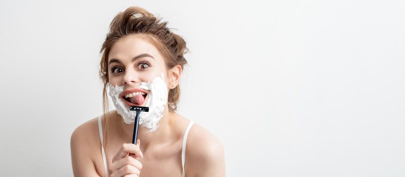 Beautiful young caucasian woman shaving her face by razor on white background. Pretty woman with shaving foam on her face