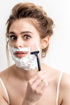 Beautiful young caucasian woman shaving her face by razor on white background. Pretty woman with shaving foam on her face