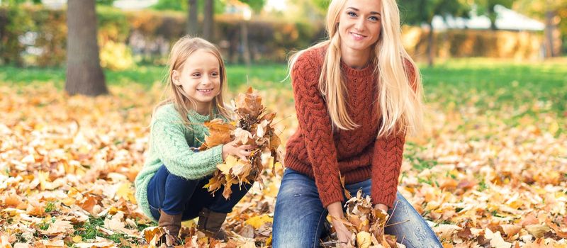 Happy young caucasian mother and little child playing with leaves in nature autumn park