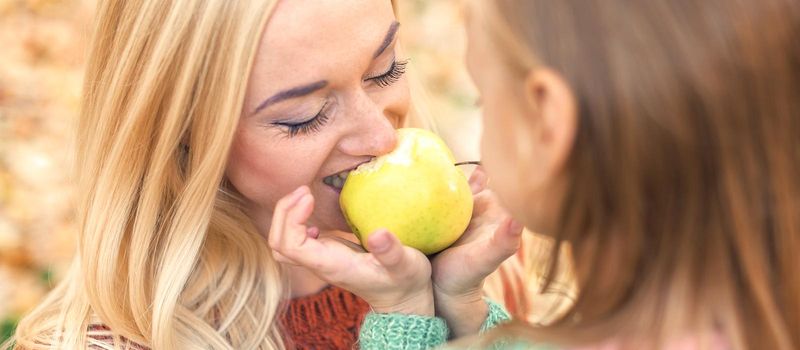 Portrait of little girl with young mother eating yellow apple in autumn park