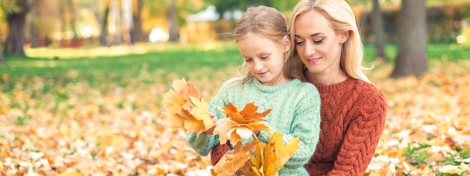 Happy young caucasian woman and little girl holding autumn yellow leaves sitting at the park