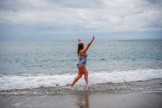 A plump woman in a bathing suit enters the water during the surf. Alone on the beach, Gray sky in the clouds, swimming in winter