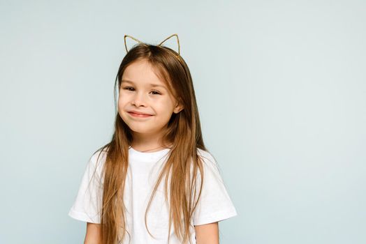 Portrait of young excited smiling girl in cat ears looking at camera on blue background with copy space.