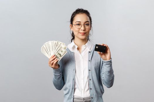 Business, finance and investment concept. Confident good-looking asian girl in glasses, female employee invest money, make good purchase or deposit, show cash dollars and credit card smile satisfied.