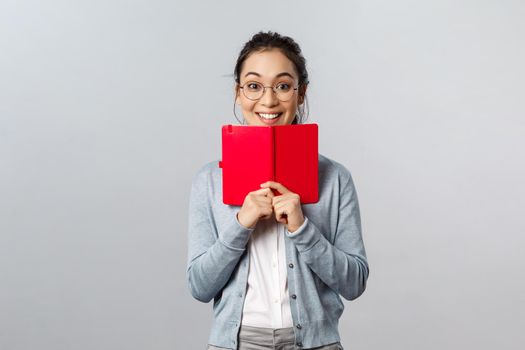 Teaching, education and university lifestyle concept. Excited happy asian woman in glasses writing down something in notebook, holding planner or secret diary pressed to chest, smiling amused.