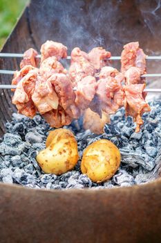 Hands of man prepares barbecue meat with potatoes on skewer by grill on fire outdoors. Concept of lifestyle rustic food preparation