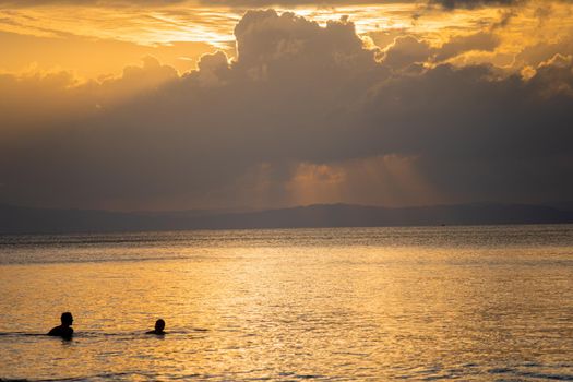 silhouette shot of people swimming under the golden sunlight god rays with clouds surrounding the sun and the blue gold water with waves lapping the shore showing peaceful life at havelock andaman islands in India