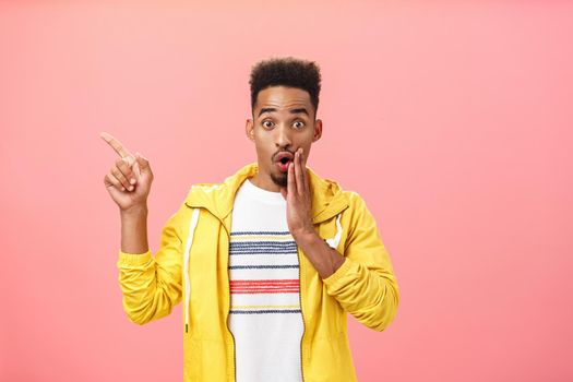 Look I am stunned. Portrait of impressed excited good-looking young male model in stylish yellow jacket opening mouth with gasp covering it with palm pointing amazed at upper left corner. Emotions and advertising concept