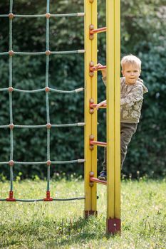 A little boy learns to climb a rope wall in a park playground. Green background for a beautiful warm summer day.