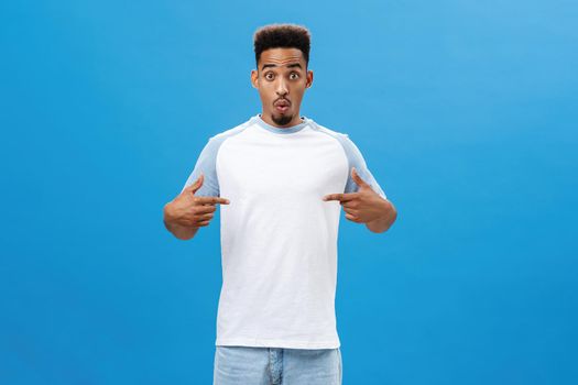Impressed and amazed good-looking young dark-skinned male model in casual t-shirt folding lips in intrigue and curiosity popping eyes at camera excited while pointing at chest over blue wall. Copy space