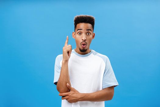 Artistic and creative dark-skinned boyfriend in trendy t-shirt folding lips in wow, raising index finger in eureka gesture making up great idea adding suggestion how solve situation over blue wall.