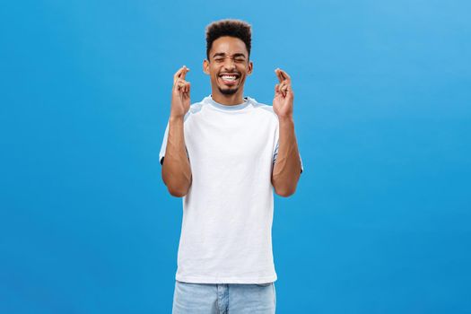 Putting all effort in pray to make dream come true. Portrait of hopeful intense and optimistic dark-skinned guy with afro haircut smiling closing eyes crossing fingers for good luck over blue wall. Body language concept