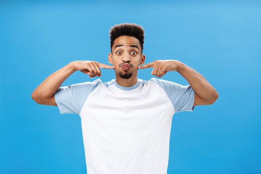 Waist-up shot of playful happy nice african american guy with tattoo on palm holding breath pouting joyfully and poking at cheeks with index fingers popping eyes while having fun over blue background. Copy space