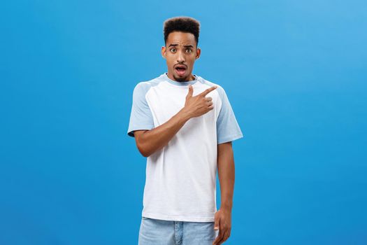 Portrait Lifestyle. Man feeling shocked finding out friend betrayed him pointing at upper right corner frowning and opening mouth with concerned upset and unhappy expression posing over blue background worried.