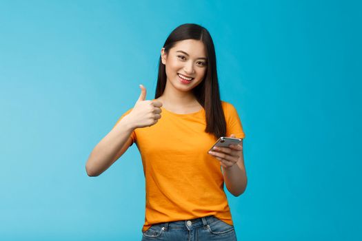 Satisfied cute asian brunette hold smartphone show thumb-up delighted, recommend awesome gadget, give approval good new app, agree friend posted good blog article, stand blue background.