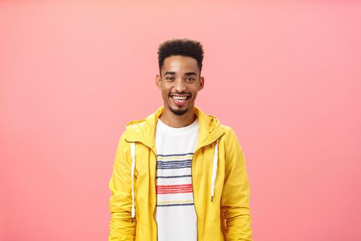 Waist-up shot of charming friendly good-looking dark-skinned male with beard and afro hairstyle in trendy yellow jacket smiling with satisfied pleased grin standing over pink background. Emotions concept