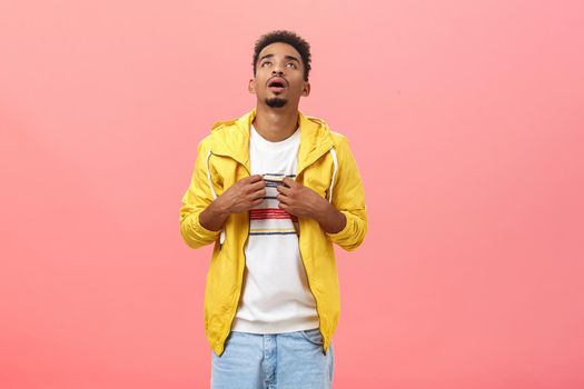 Stylish dark-skinned man suffering from hot weather breathing out waving with t-shirt to cool standing in discomfort over pink background wearing inappropriate clothes for sunny summer day. Copy space