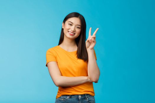 Optimistic cute tender asian young millennial girl send positive vibes, tilt head show peace, victory sign, smiling broadly, wish fantastic holidays, stand blue background joyful upbeat.