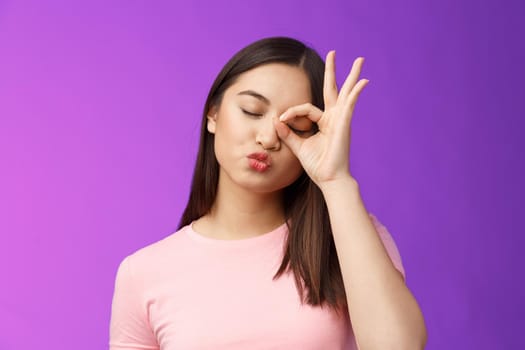 Cute silly asian female brunette folding lips kiss lovely expression, close eyes dreamy send muah, show perfection, okay ok sign, stand purple background carefree and cheerful. Copy space