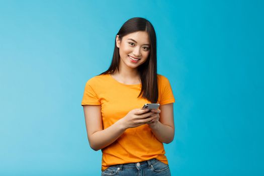 Cheerful friendly modern young asian girl student messaging friend, hold smartphone texting followers, using new internet application, try out app stand satisfied smiling camera, blue background.