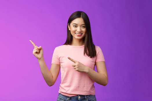 Friendly cute pleasant asian woman showing way, indicating interesting promo, pointing left smiling broadly, introduce advertisement product, stand purple background cheerful. Copy space