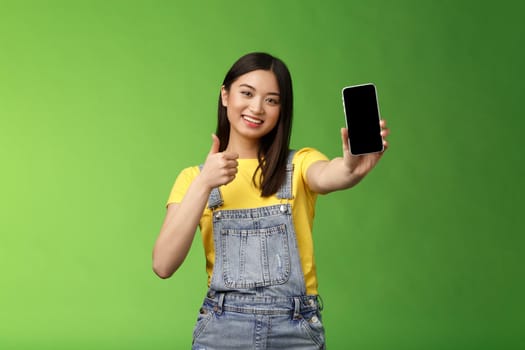 Cheerful cute asian woman judging good smartphone game, show thumb-up like sign, extend arm hold telephone screen, present awesome app, promoting application, stand green background.