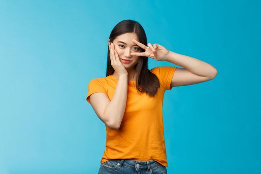 Sassy stylish good-looking asian assertive korean girl show peace victory sign on eye, touch cheek look empowered and motivated, standing cheeky coquettish blue background, wear yellow t-shirt.