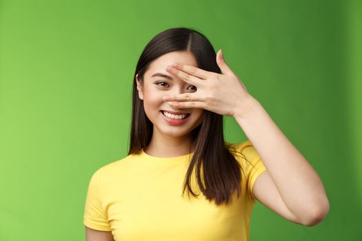 Cheerful enthusiastic cute asian brunette girl smiling broadly, toothy positive grin, cover eyes peeking through fingers amused, fascinated expression, stand green background. Copy space