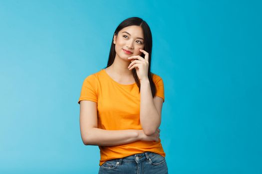 Joyful confident asian girl with dark haircut tilt head, dreaming, imaging interesting plan, smiling cunning, have idea, thinking and making choice, look up thoughtful, stand blue background.