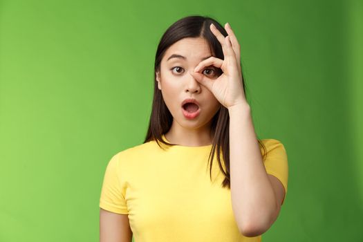 Amazed speechless cute asian young girl drop jaw astonished, look through ok circle sign, drop jaw speechless, impressed look camera, check out incredible promo, stand green background.