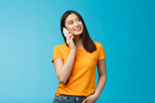Friendly cute urban asian girl look up dreamy relaxed, smiling joyfully hold smartphone near ear, talking casually friend via telephone, satisfied good mobile connection, stand blue background.