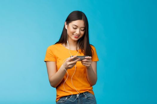 Happy entertained asian cute girl brunette, wear earphones not disturbed people in cafe while playing favorite smartphone game, hold phone horizontal, smiling look enthusiastic cellphone screen.