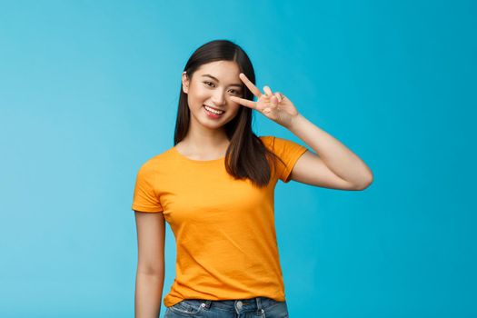 Friendly cheerful asian girlfriend look joy happiness, show peace victory sign, smiling toothy positive, enjoy lucky perfect summer sunny day, stand blue background carefree wear yellow t-shirt.