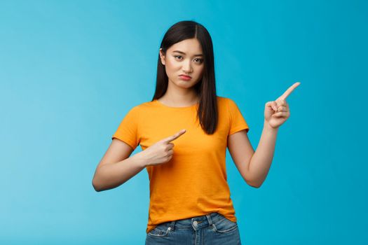 Picky disappointed asian girl with unsatisfied face, pointing upper right corner, sulking offended, feel jealous friend went summer vacation abroad alone, stand blue background displeased.