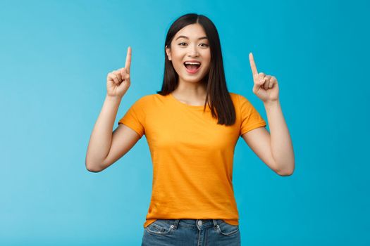 Amused asian girl introduce new product sharing promo with you, raise hands pointing fingers up smiling broadly, look excited and upbeat, enthusiastic advertising, stand blue background.