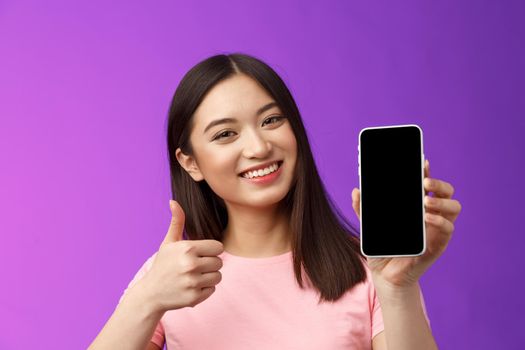Close-up satisfied happy attractive modern asian woman give positive feedback, show smartphone display, introduce good app, raise thumb-up like application, stand purple background delighted.