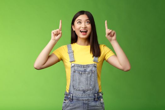 Carefree amused asian brunette in yellow t-shirt dungarees, smiling amazed and fascinated, look pointing up, observe cool new promo, grinning excited, enjoy watching top copy space.