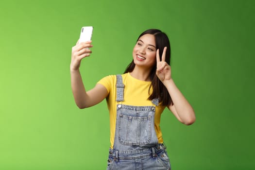 Tender cute asian brunette female, hold smartphone, posing selfie, look telephone screen, photographing, show victory peace sign, joyfully chatting friend abroad, stand green background. Copy space