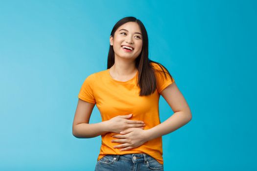 Happy staffed cute asian girl feelings satisfaction after eating favorite food, smiling broadly rubbing tummy, touch stomach from delight and joy, like eat delicious meals, stand blue background.