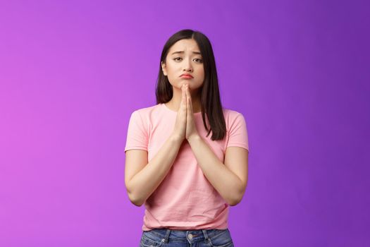 Gloomy cute asian daughter begging mother buy new phone, supplicating, hold hands pray, sulking frowning pretty please, asking apology, pleading favor, stand purple background. Copy space