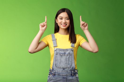 Friendly cute good-looking asian brunette female pointing up, raise fingers top advertisement, self-assured showing promo, smiling satisfied, introduce new product, green background.