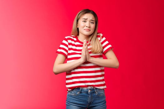 Gloomy upset girl in need begging mercy, asking apology feel guilty sad. Clingy cute daughter frowning grimacing unhappy sorrow hold hands pray pleading borrow car, supplicating red background.