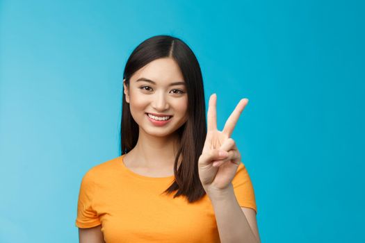 Cheerful friendly asian woman in yellow t-shirt smiling broadly, show number two, twice, win second place, smiling joyfully, make take-away order, book sits, stand blue background.