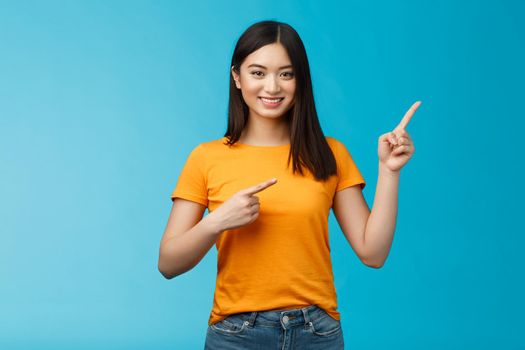 Friendly cheerful asian brunette girl inviting visit link interesting place, pointing upper right corner smiling joyfully camera, telling you about good promotion, stand blue background upbeat.